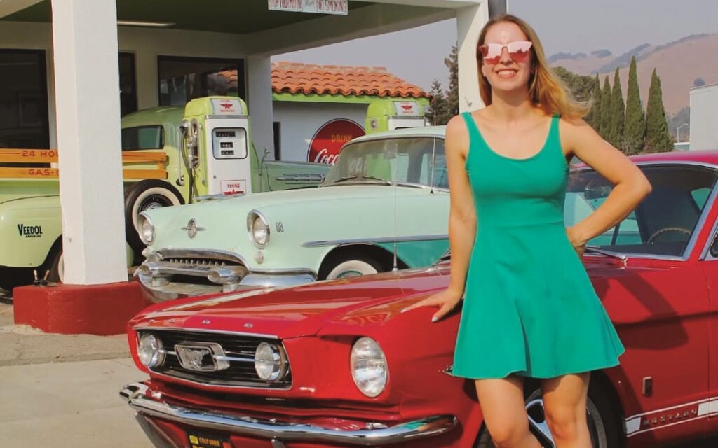 Francesca devant sa Mustang GT 1966 dans une station-service vintage